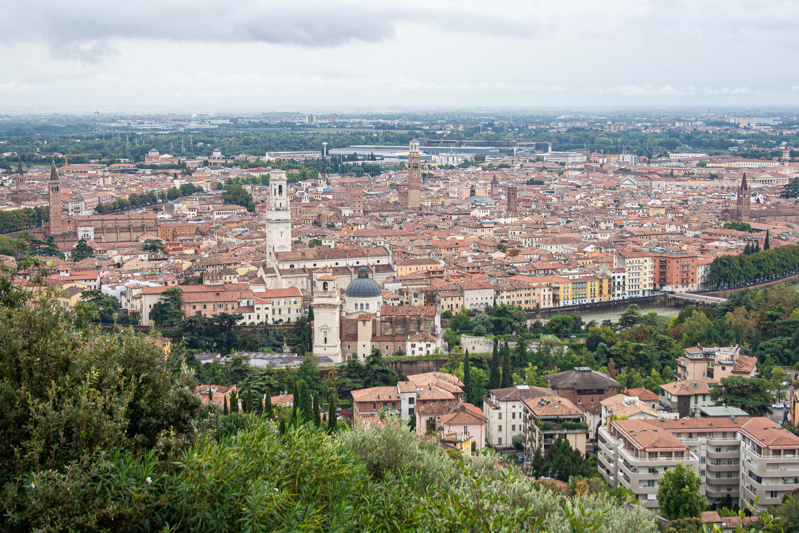 Das Bild zeigt einen Blick auf die Altstadt von Verona, Italien. Zu sehen sind historische Gebäude wie die Basilica di Santa Anastasia, die Cattedrale di Verona (Duomo di Verona) und der markante Torre dei Lamberti. Die typisch orangefarbenen Dächer und der Fluss Etsch prägen das Stadtbild.