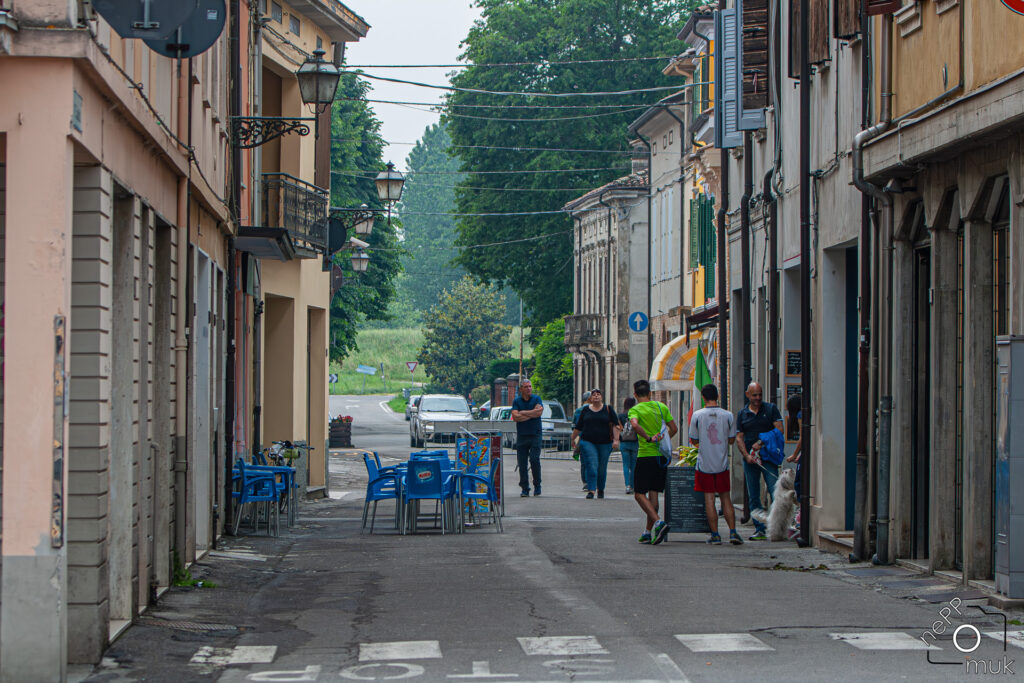Die Via Felice Cavallotti, von der Piazza aus fotografiert. Die Straße ist flankiert von historischen Gebäuden, blauen Café-Stühlen und Passanten, die durch die Gasse schlendern. Im Hintergrund öffnet sich die Landschaft mit Bäumen und einem entfernten Weg.